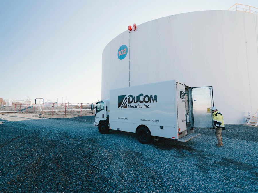 A DuCom Electric box truck parked on gravel beside a large white storage tank, with a technician in hi-vis at the open rear door