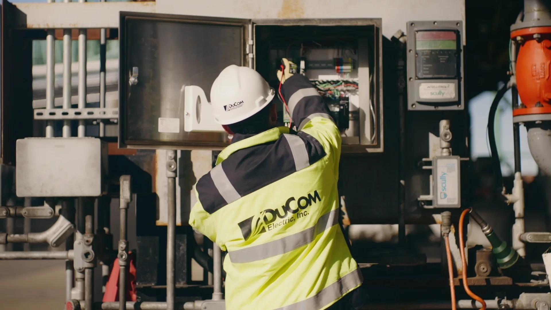 A DuCom technician in a hard hat and hi-vis jacket works inside an open control enclosure among valves and meters