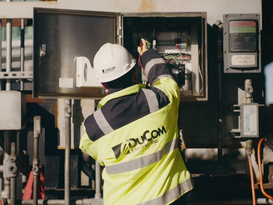 A DuCom technician in a hard hat and hi-vis jacket works inside an open control enclosure among valves and meters