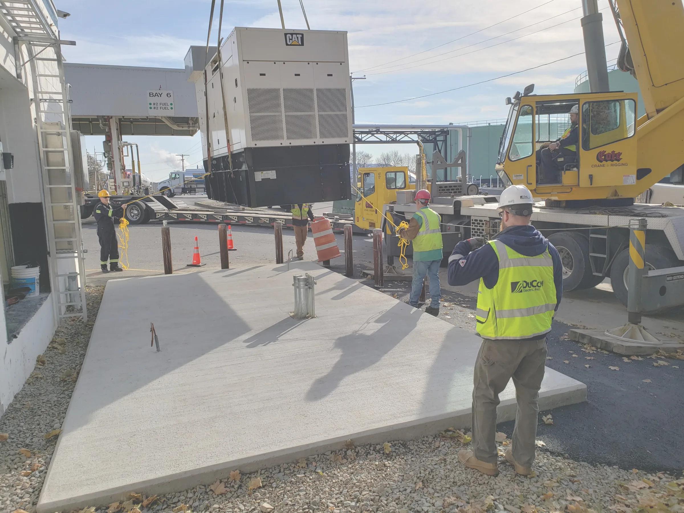 A crane lowers a generator onto a new concrete pad at a fuel loading rack, guided by crew members in DuCom hi-vis