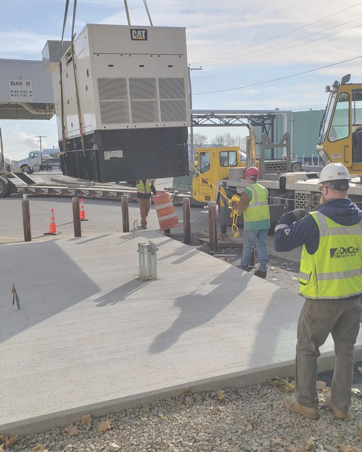 A crane lowers a generator onto a new concrete pad at a fuel loading rack, guided by crew members in DuCom hi-vis