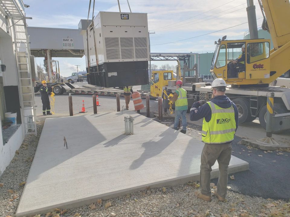 A crane lowers a generator onto a new concrete pad at a fuel loading rack, guided by crew members in DuCom hi-vis
