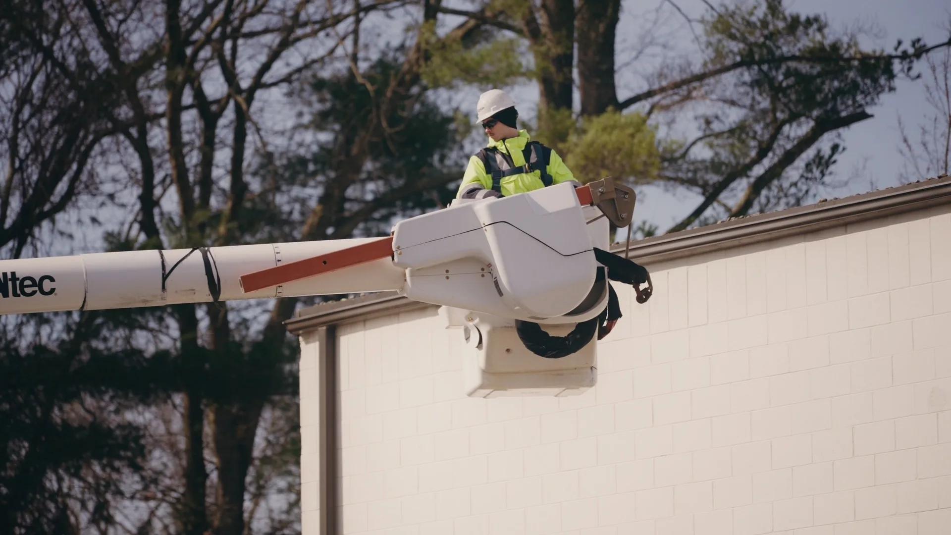 A technician in a hard hat and hi-vis jacket works from a bucket lift at the roofline of a block building