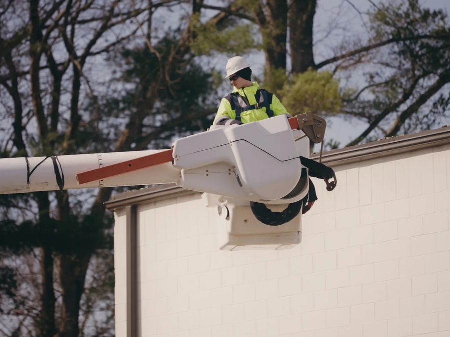 A technician in a hard hat and hi-vis jacket works from a bucket lift at the roofline of a block building