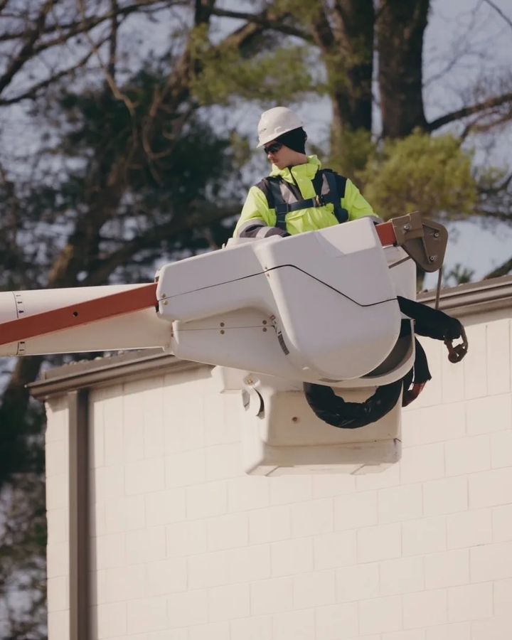 A technician in a hard hat and hi-vis jacket works from a bucket lift at the roofline of a block building