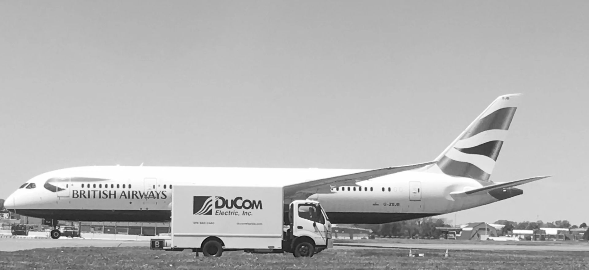 A DuCom Electric truck on an airport apron beside a parked airliner, in black and white