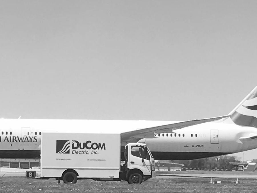 A DuCom Electric truck on an airport apron beside a parked airliner, in black and white