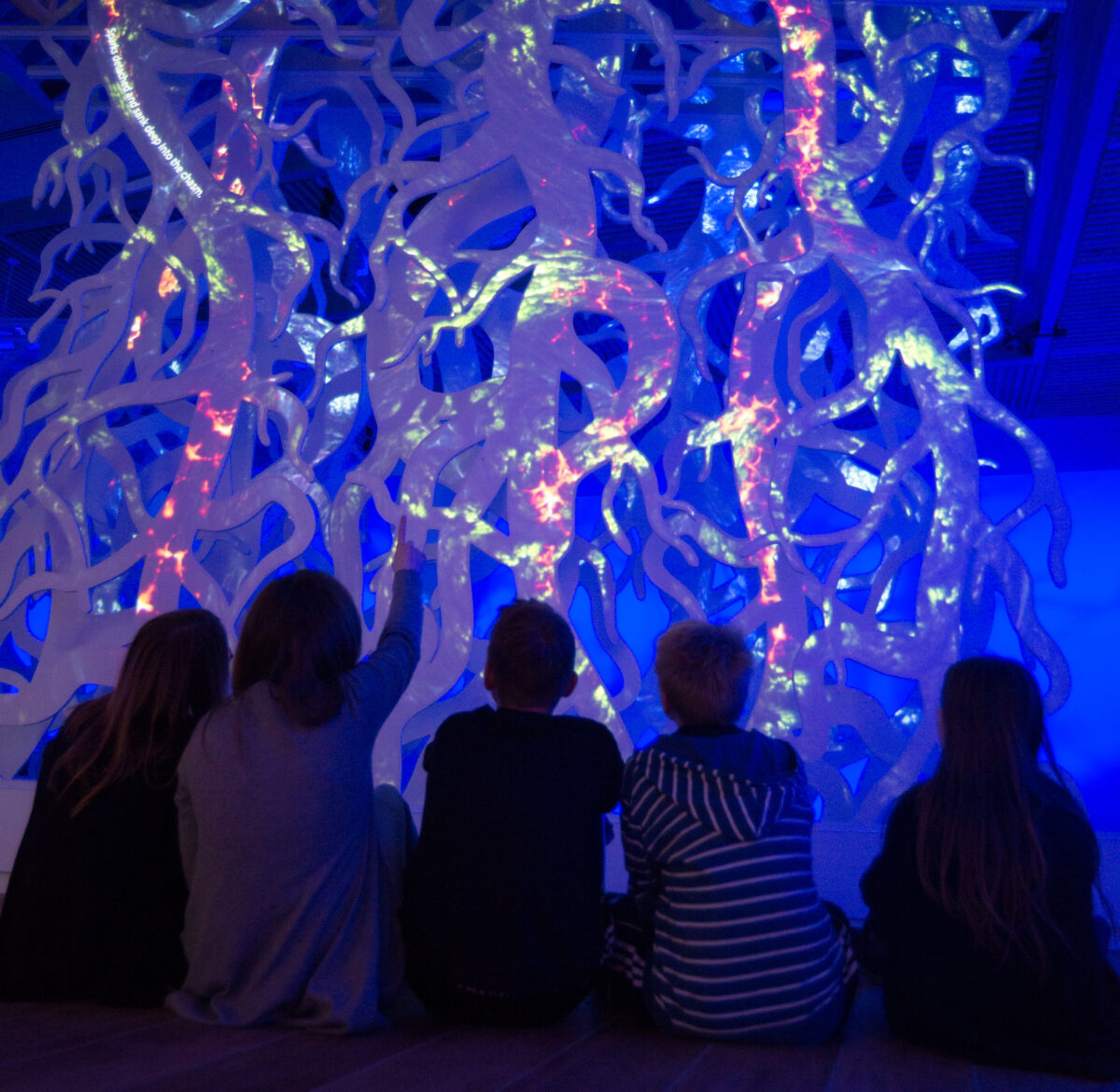 Five children sit in front of a sculpture of Yggdrasil's roots with light projected onto it.