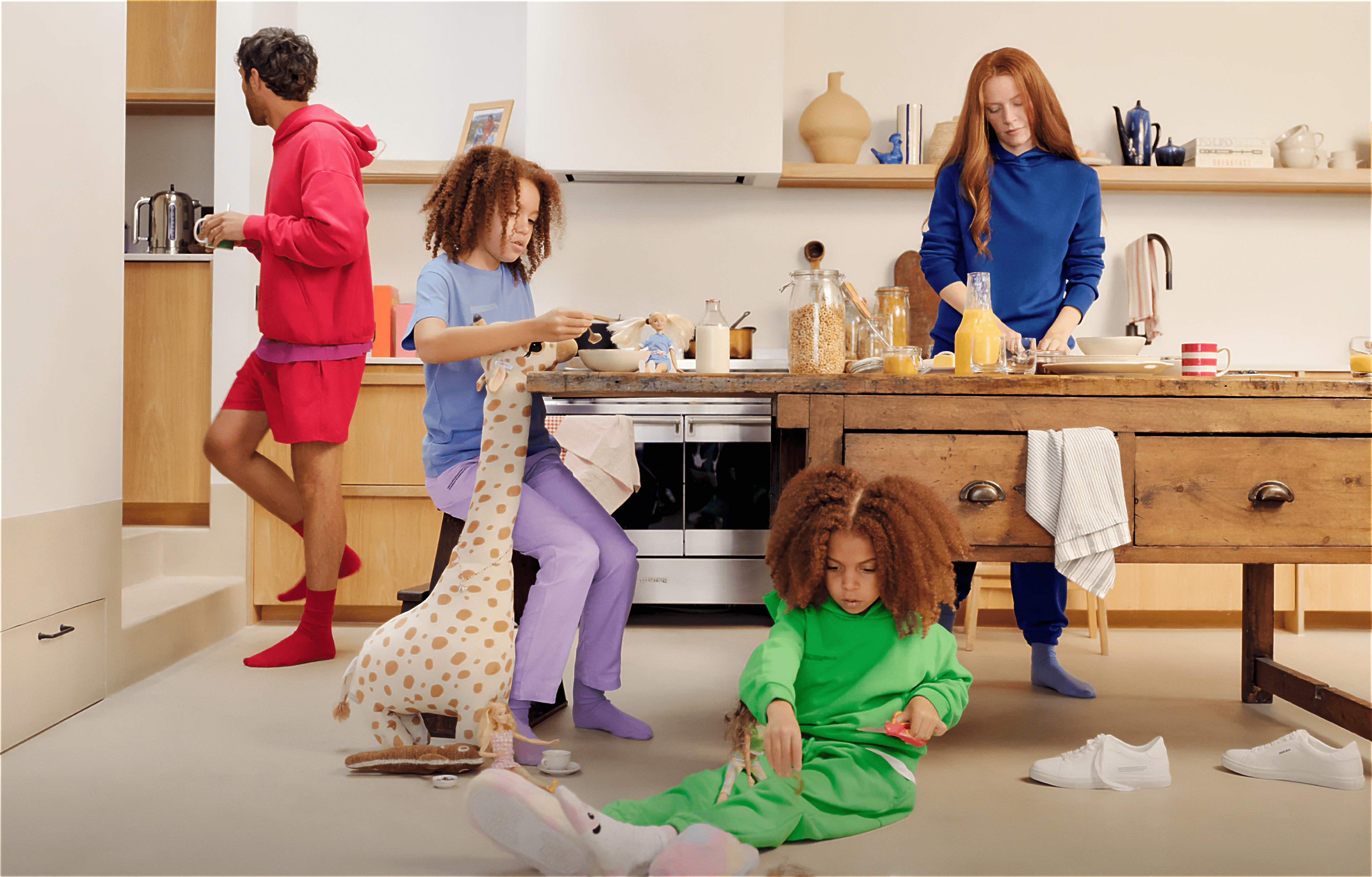A cozy kitchen scene featuring a family wearing colorful, sustainable loungewear by Pangaia. The father, dressed in a red hoodie and shorts, prepares a drink at a counter. Two children play nearby: one seated on a stool in purple pants and a blue shirt, holding a stuffed giraffe toy, while the other, in a green outfit, plays with toys on the floor. A woman in a blue outfit stands at the kitchen island, preparing breakfast with orange juice and cereal jars on the countertop. The scene is warm and inviting, emphasizing a relaxed, eco-friendly lifestyle.