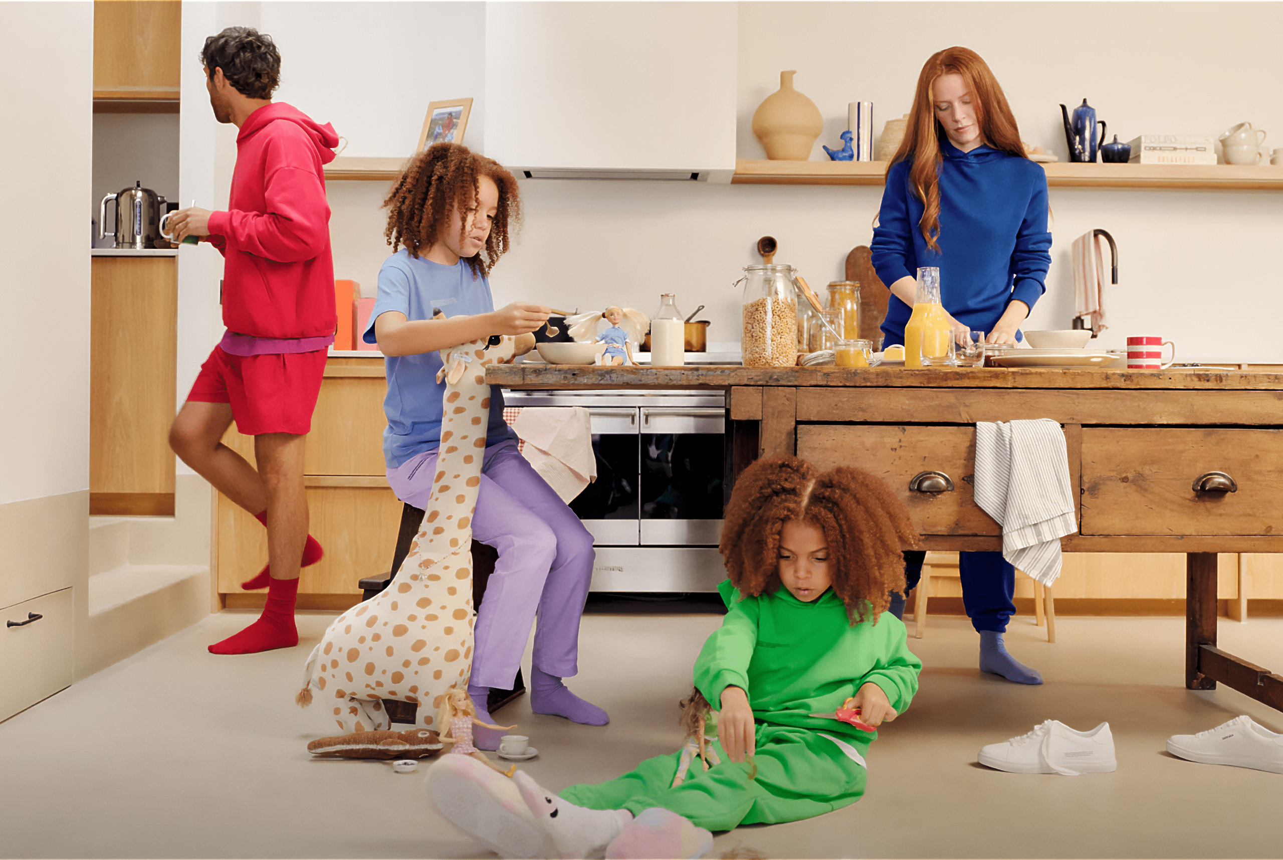 A cozy kitchen scene featuring a family wearing colorful, sustainable loungewear by Pangaia. The father, dressed in a red hoodie and shorts, prepares a drink at a counter. Two children play nearby: one seated on a stool in purple pants and a blue shirt, holding a stuffed giraffe toy, while the other, in a green outfit, plays with toys on the floor. A woman in a blue outfit stands at the kitchen island, preparing breakfast with orange juice and cereal jars on the countertop. The scene is warm and inviting, emphasizing a relaxed, eco-friendly lifestyle.