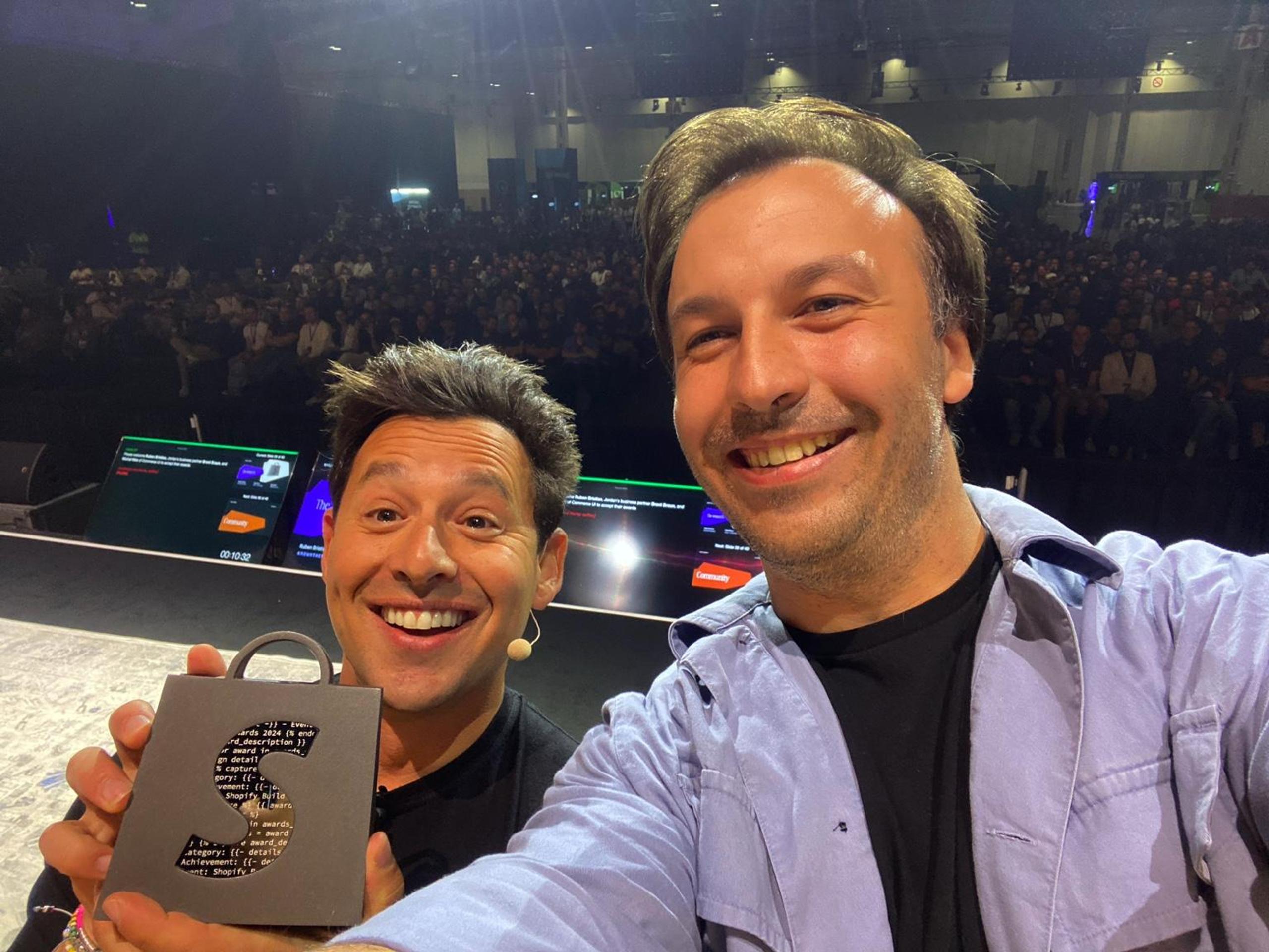 Harley Finkelstein, President of Shopify, and Michal Klim, Co-Founder and CTO at Commerce-UI, smiling and posing for a selfie on stage at an event. Harley is holding a Shopify Build Award figurine in the shape of a gray shopping bag with a large 'S' on it, containing code snippets inside the letter. The background shows a large audience and stage monitors.