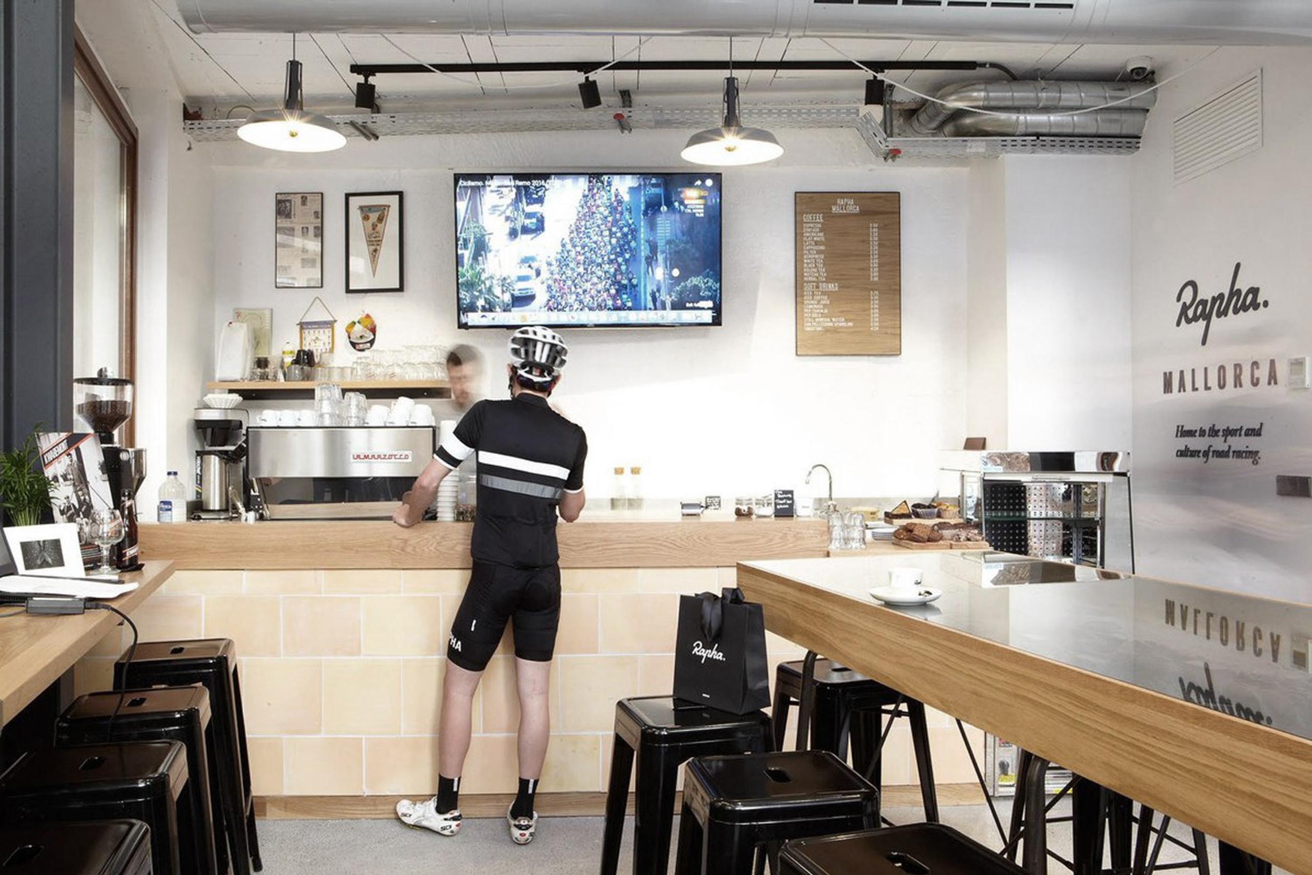 A man with a bike helmet stands in a Rapha Clubhose Mallorca, indicating a moment of ordering in a cafe.