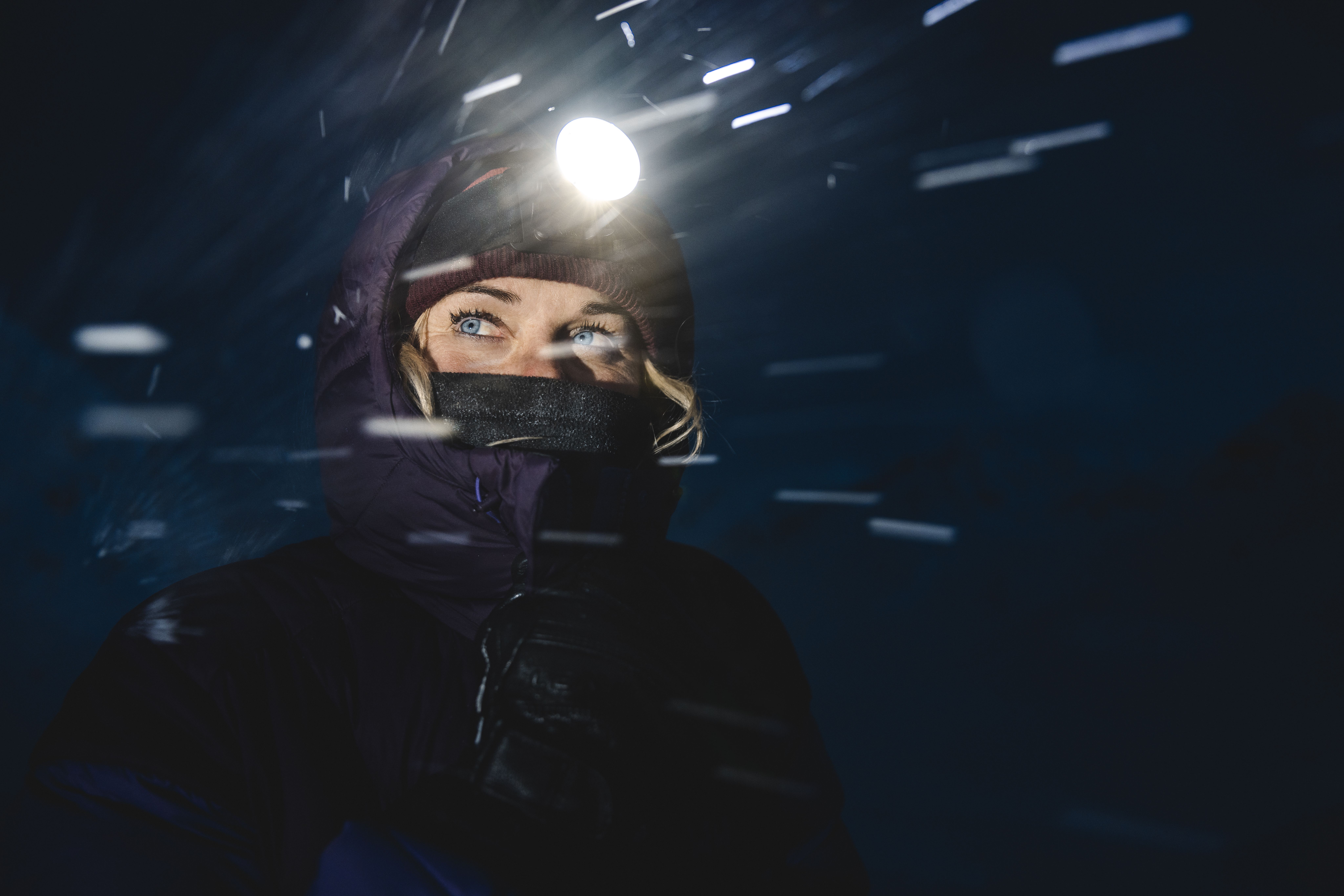 Close-up of a bundled-up hiker in blowing snow at night, wearing a bright Lupine headlamp that casts streaks of light across the dark scene.