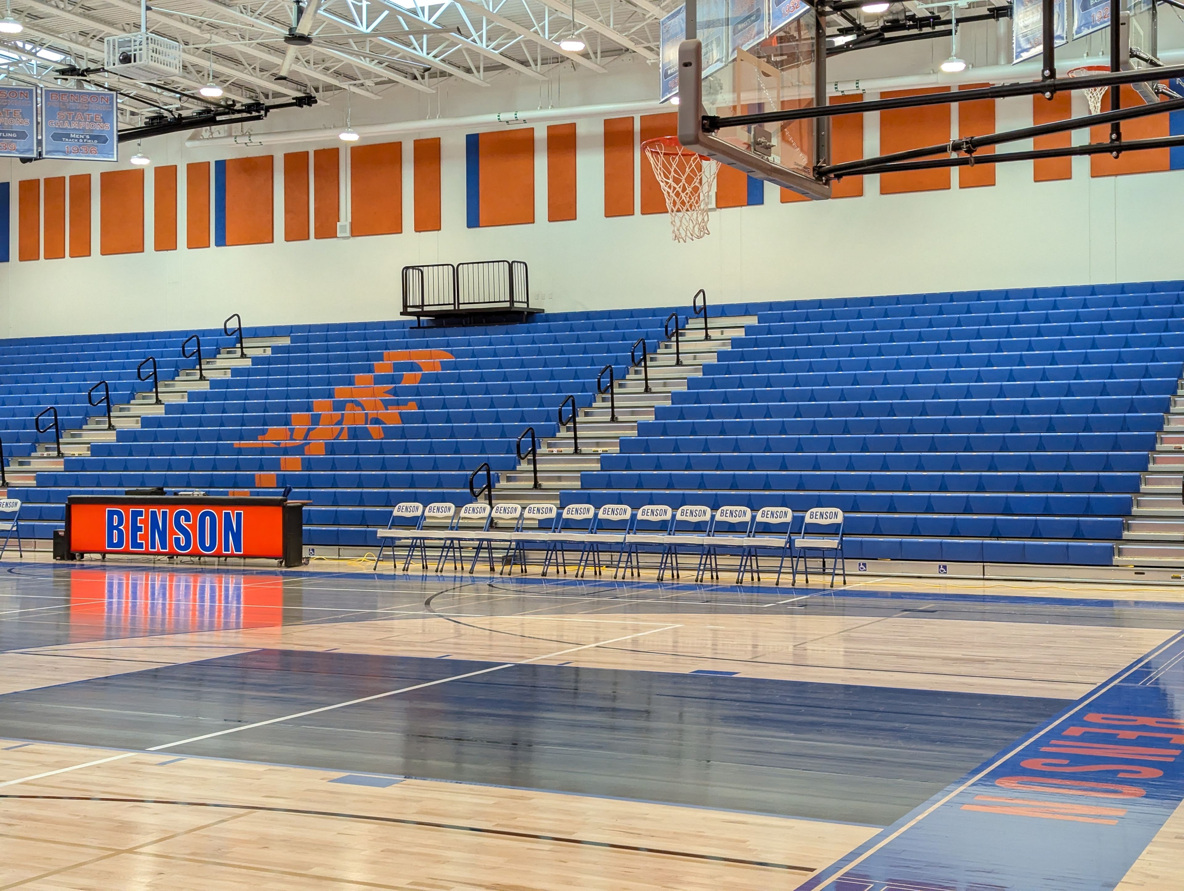 Benson gym, with blue bleachers that have an orange "BT" logo on them, a desk that says "Benson", and courtside chairs.