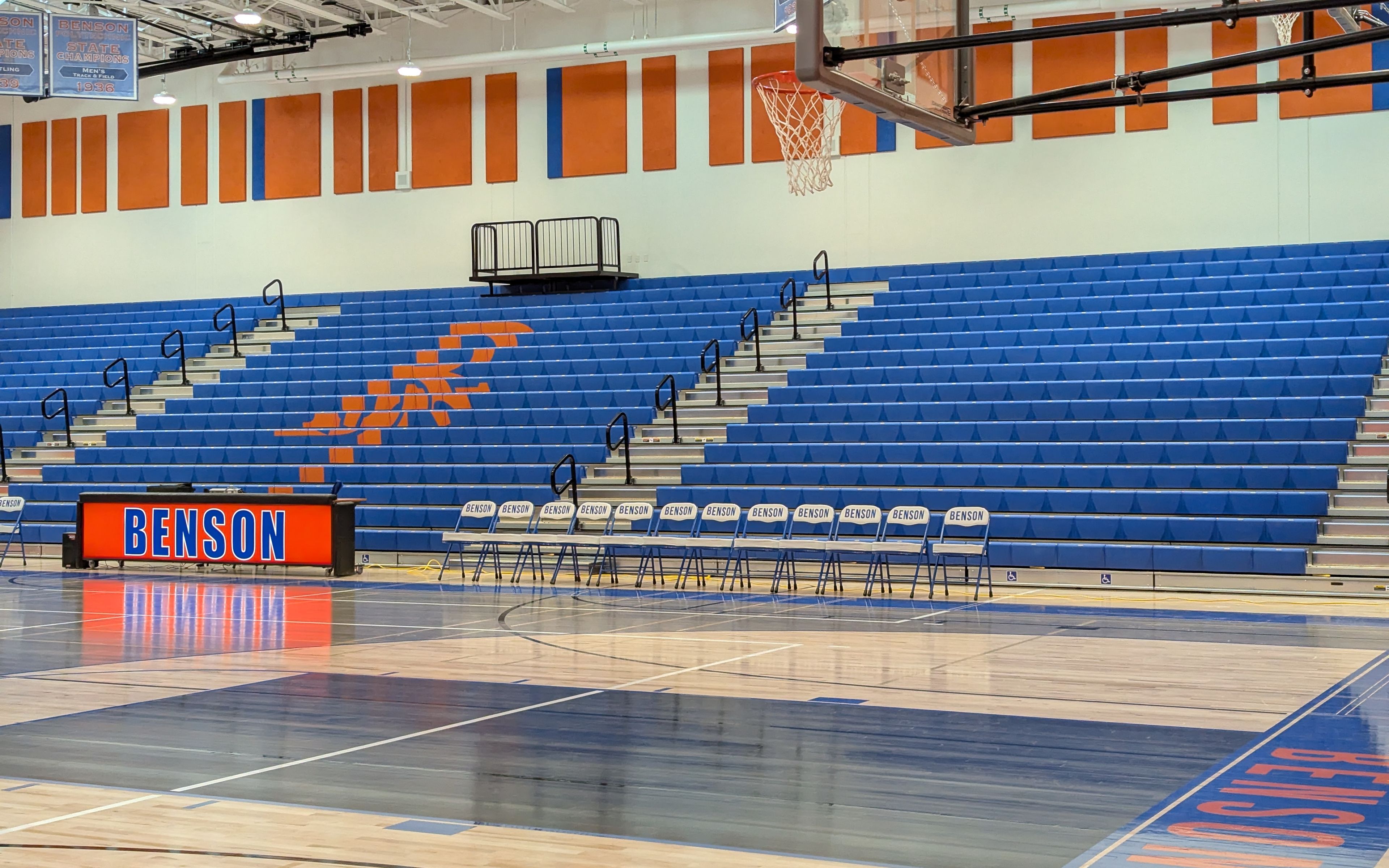 Benson gym, with blue bleachers that have an orange "BT" logo on them, a desk that says "Benson", and courtside chairs.