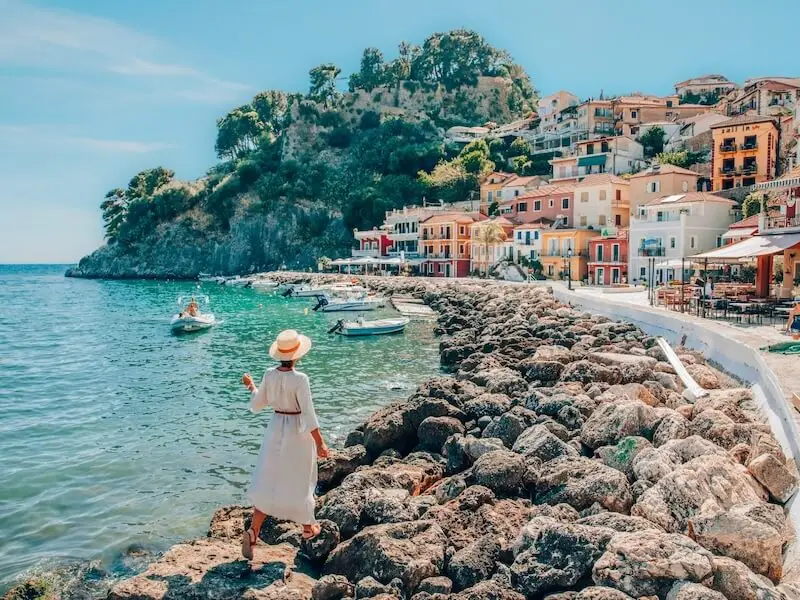 Woman walking along the colourful seafront of Parga, Greece, overlooking boats and pastel hillside villas — a picturesque stop for luxury travellers and Ionian catamaran cruises with Yacht Getaways.
