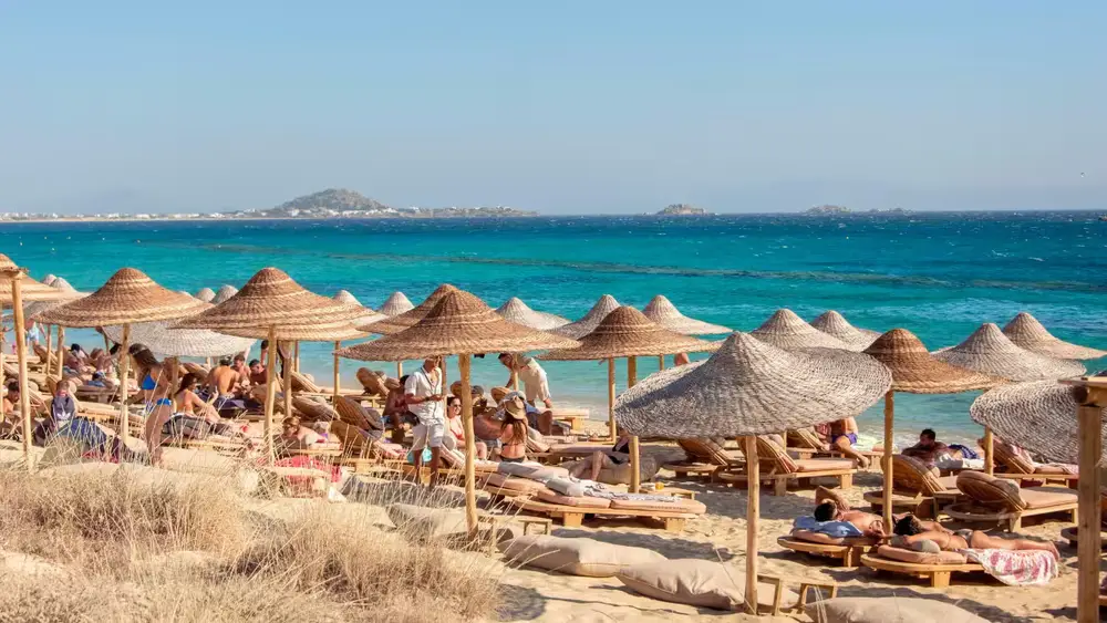 Seaside beach club with sun loungers and straw parasols beside turquoise water on a Cyclades island holiday in Greece.