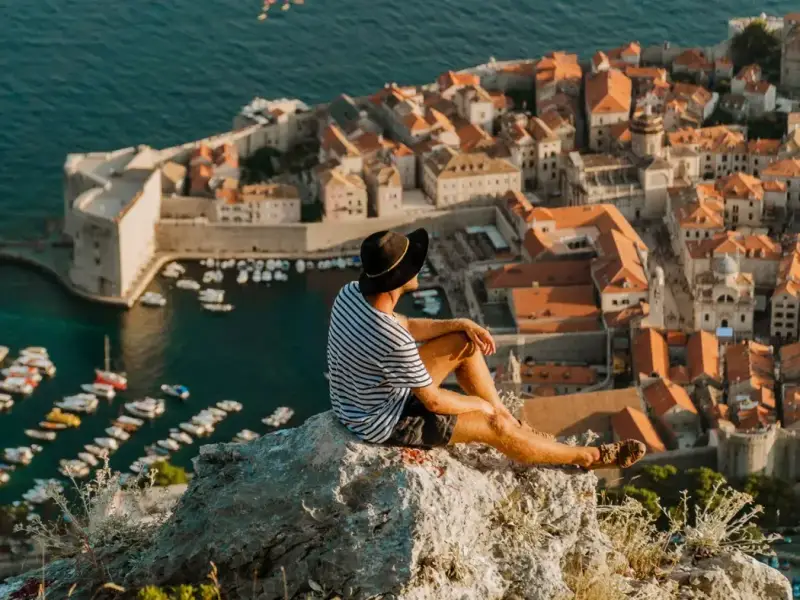 Traveller sitting on a rocky viewpoint above Dubrovnik Old Town, looking over the stone walls, terracotta rooftops and harbour boats on Croatia’s Adriatic coast — a signature stop for anyone planning the best places to visit in Croatia in 2026, and a natural fit for Yacht Getaways-style island and coastal routes.