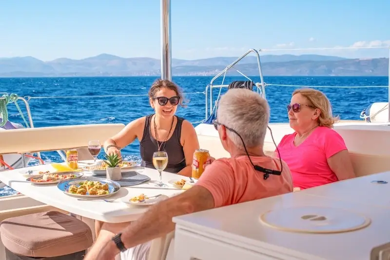 A group of luxury travellers laughing and enjoying a drink on the back of a premium catamaran while cruising the Mediterranean.