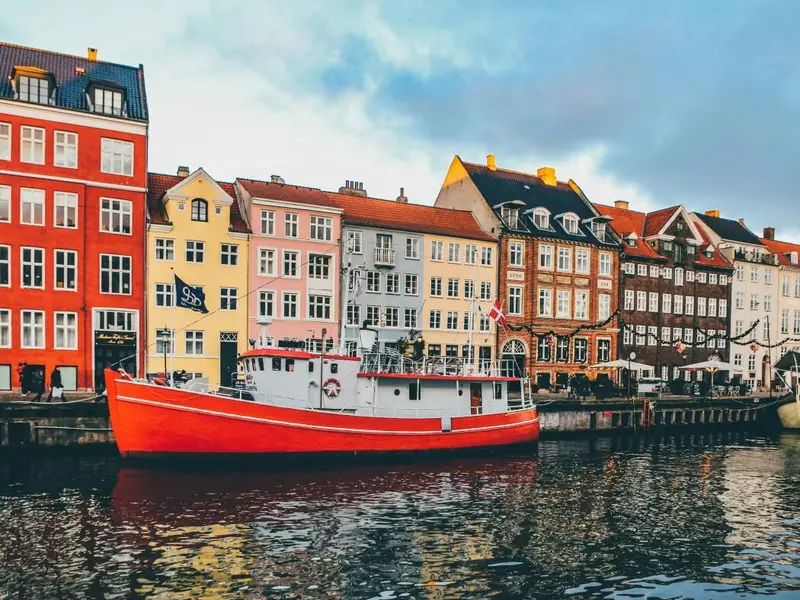 A bright red boat sits on the canal in Copenhagen’s Nyhavn, Denmark, backed by colourful historic townhouses reflected in the water under a moody sky. A classic Copenhagen waterfront scene that fits an article on the safest places to visit in Europe, highlighting Denmark as a relaxed, well-organised destination with excellent public transport, walkable neighbourhoods, and an easy, comfortable city-break feel for mature travellers.