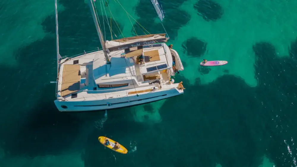Aerial view of a premium sailing catamaran anchored in crystal-clear turquoise Mediterranean waters, with guests paddleboarding nearby during a Yacht Getaways sailing holiday.
