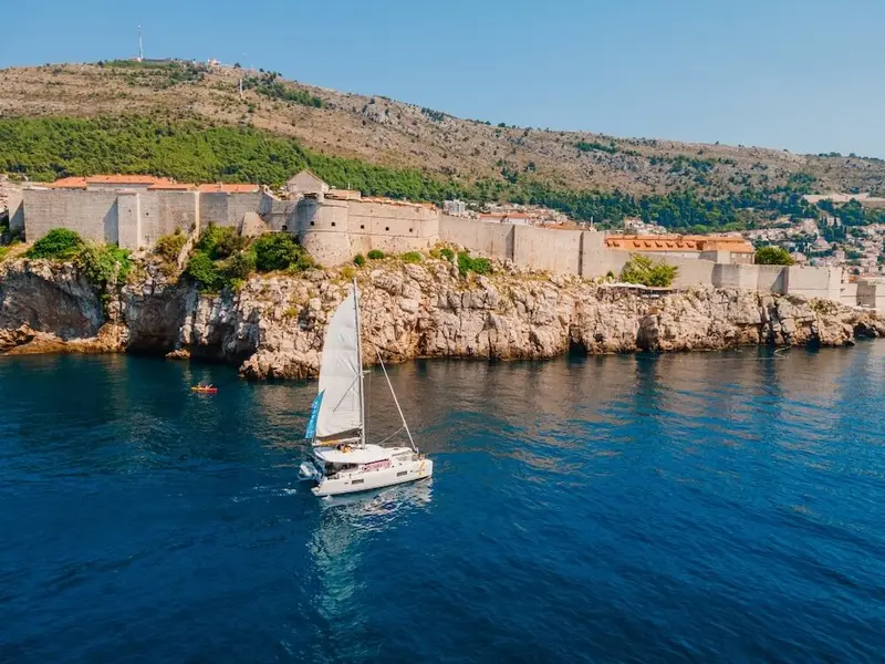 A catamaran sails across calm, deep-blue water below the historic stone city walls and fortifications of Dubrovnik, Croatia, with green hills rising behind the Old Town. A peaceful Adriatic coastline scene that fits an article on the safest places to visit in Europe, highlighting Croatia as a relaxed Mediterranean destination known for its walkable historic cities, scenic coastal views, and easy-going travel atmosphere.