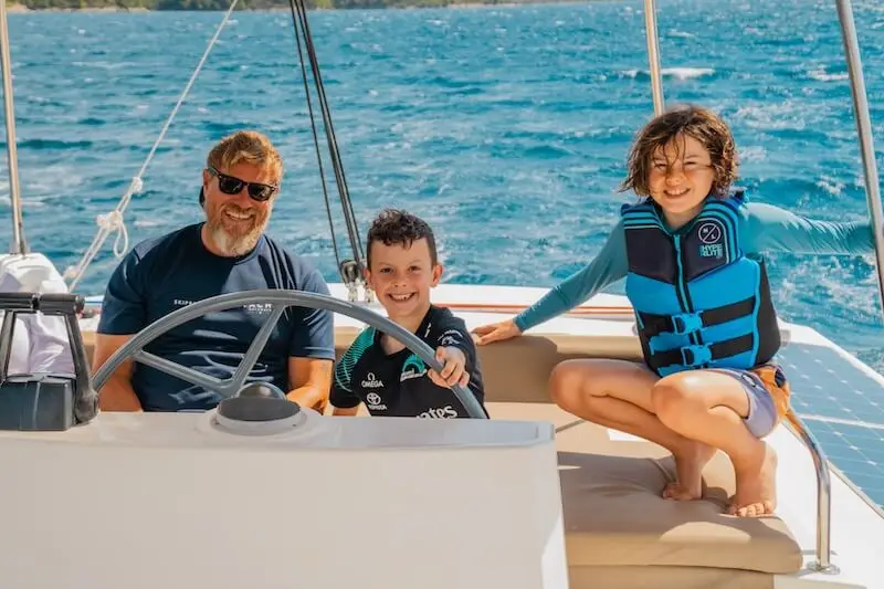 Two happy kids sit with a captain on a family cruise aboard a luxury catamaran in the Mediterranean.