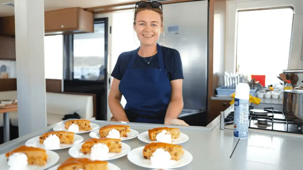 Smiling Yacht Getaways host standing in the galley of a premium catamaran beside freshly prepared individual desserts topped with cream, ready to serve to guests during their sailing holiday.