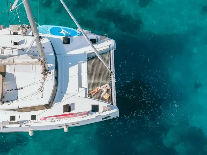 uest relaxing on the catamaran net above clear turquoise Mediterranean water on a Yacht Getaways sailing holiday, celebrating our B Corp Certified responsible luxury travel.