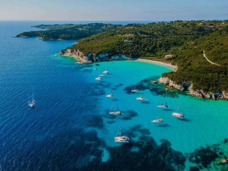 High aerial view of Voutoumi Beach on Antipaxos, with brilliant turquoise water, anchored yachts, and a pine-covered coastline leading down to the bay. Often considered one of the most beautiful beaches in the Ionian Islands, Voutoumi is a popular stop on sailing holidays in Greece for its Caribbean-like colours and clear swimming water.