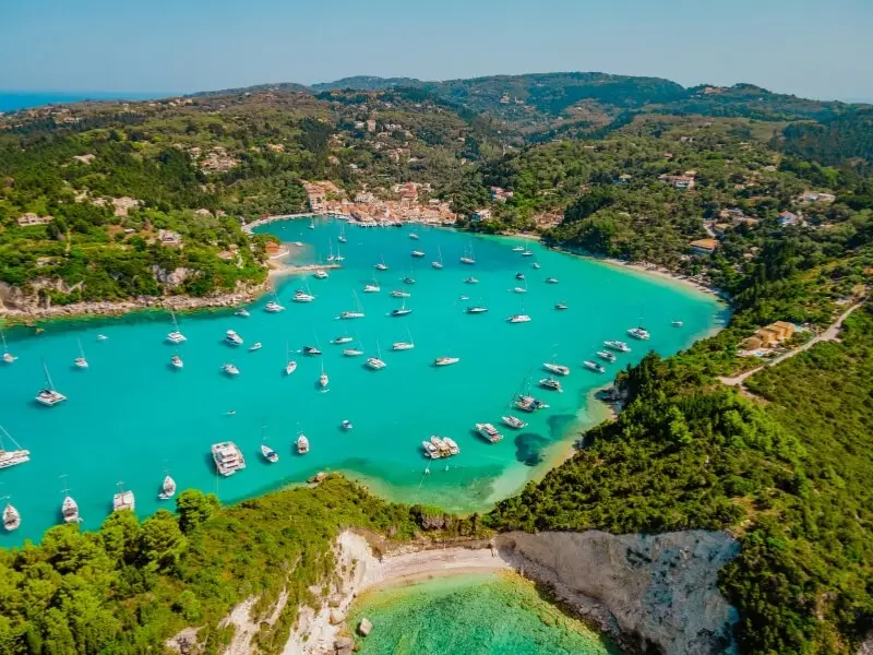 Aerial view of Lakka, Paxos, a sheltered Ionian bay in Greece with yachts at anchor and a harbour village surrounded by green hills.