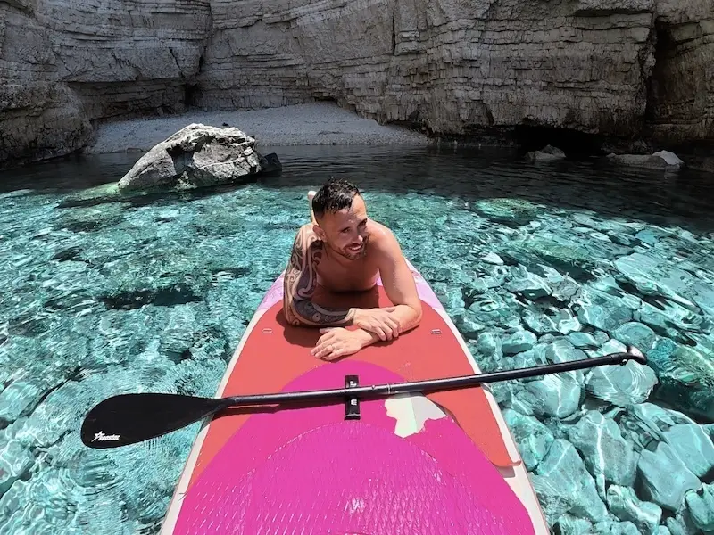 A man relaxes and smiles on a paddleboard floating atop bright blue Mediterranean waters while on a catamaran cruise.