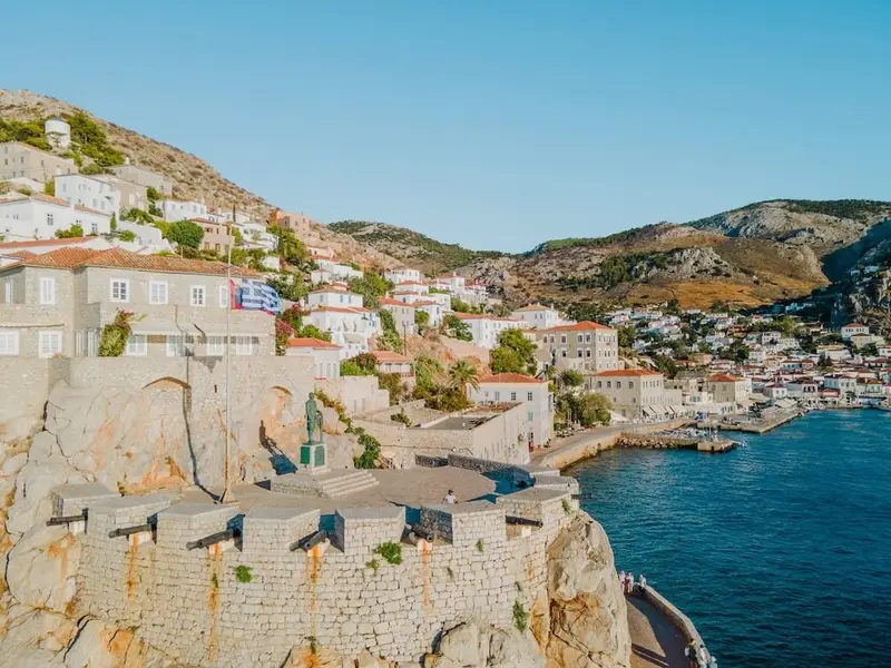Hydra Island harbour in Greece, showing the stone waterfront fortifications with old cannons, the Greek flag flying above whitewashed houses on the hillside, and the deep-blue sea of the Saronic Gulf — a classic stop on an Athens island-hopping sailing route.