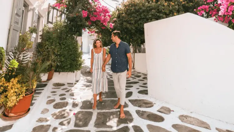 Couple holding hands while exploring a picturesque Greek island village, walking along a traditional cobblestone street lined with whitewashed buildings, colourful bougainvillea and Mediterranean greenery.