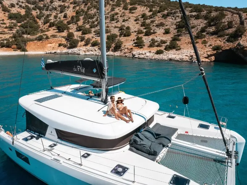A luxury catamaran anchored in a beautiful blue bay in Greece while a couple sits on top of the deck sunbathing during their luxury sailing holiday in the Greek Islands.