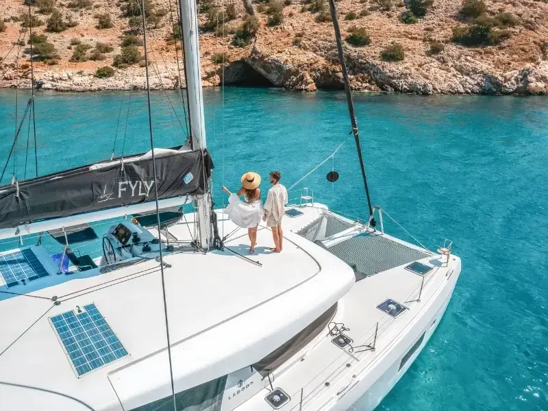 A luxury catamaran anchored in a bright blue bay in the Greek Cyclades islands, with a couple standing atop looking out at a sea cave.