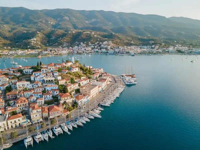 Aerial view of Poros Island harbour in Greece, with colourful waterfront houses, a marina lined with sailing yachts, and calm blue water backed by green hills across the Saronic Gulf — an iconic stop on an Athens island-hopping sailing holiday.