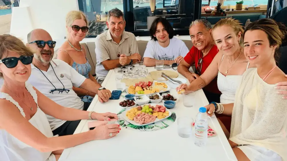 Group of adult guests smiling around a dining table aboard a Yacht Getaways catamaran, sharing a Mediterranean-style meal in the shaded outdoor dining area.