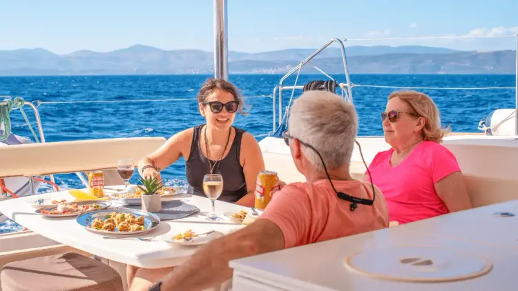 Yacht Getaways guests sharing a relaxed lunch with food and drinks in the outdoor dining area of a premium sailing catamaran, enjoying good company and Mediterranean sea views.