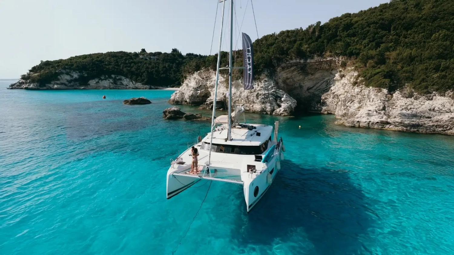 Luxury catamaran anchored in a bright turquoise bay surrounded by rugged coastline with a couple on the bow enjoying a cruise book on travel deals with Yacht Getaways.