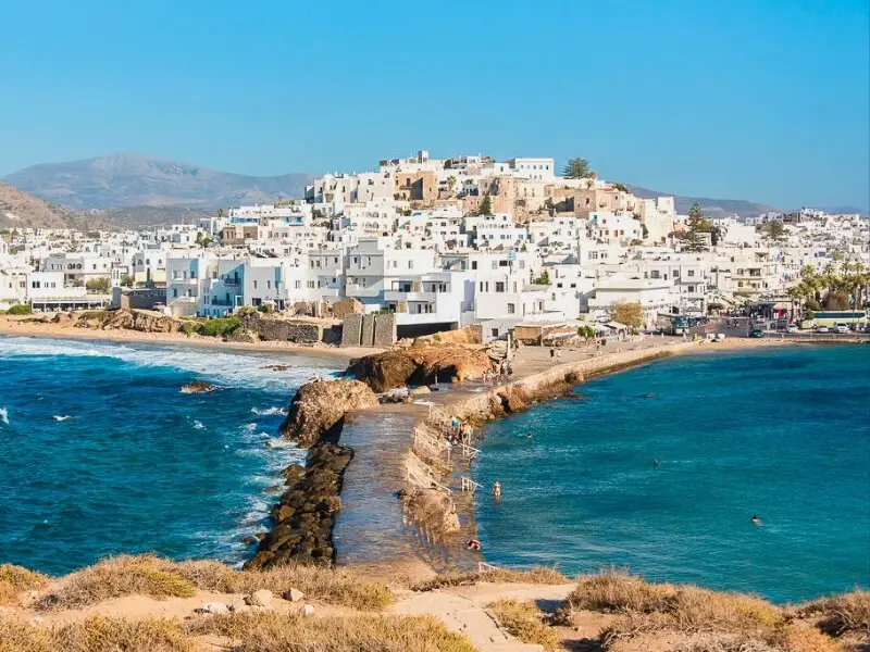 Whitewashed Cyclades town of Naxos by the sea with a stone causeway and swimmers, Greek Islands, on a luxury sailing holiday with Yacht Getaways.
