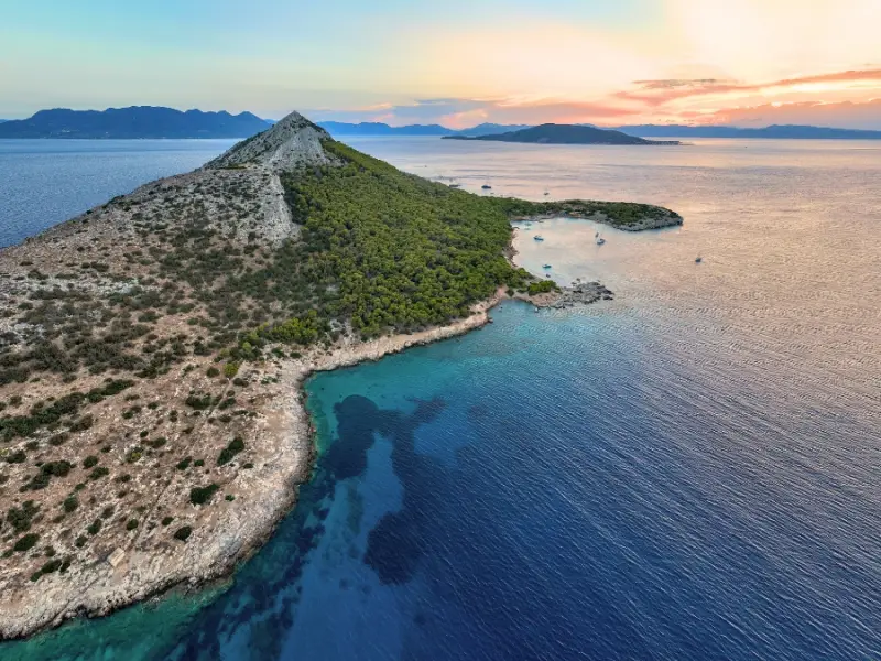 Drone view of Moni Island near Aegina in the Saronic Gulf, showing its distinctive pointed hillside, peaceful coves, and calm sea at sunset. This uninhabited island is a favourite stop on Greece sailing itineraries thanks to its natural beauty, quiet anchorages, and feeling of complete escape just a short sail from Athens.