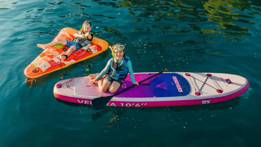 Two children wearing life jackets and snorkelling masks enjoying watersports on a family sailing holiday, with one sitting on a paddleboard and the other relaxing on an inflatable raft in clear Mediterranean waters.