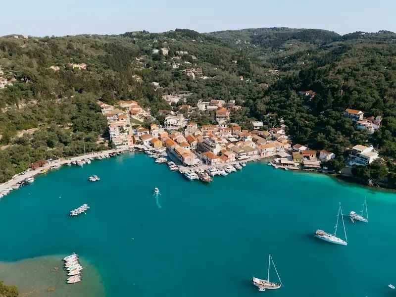 Aerial drone shot of a small coastal Greek island town with sailing yachts anchored in a turquoise bay.