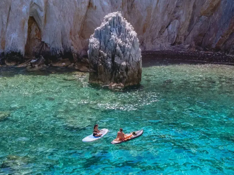 Travellers paddleboarding in clear turquoise Mediterranean water beside dramatic rocky cliffs, showing the calm and active side of blue health travel. This image reflects how a luxury yacht holiday in Greece or Croatia can combine gentle movement, time in nature and peaceful sea views for a more restorative wellness escape.