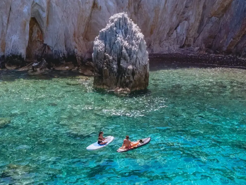 A couple paddle board in bright blue waters in a bay in the Mediterranean while on a luxury sailing holiday.