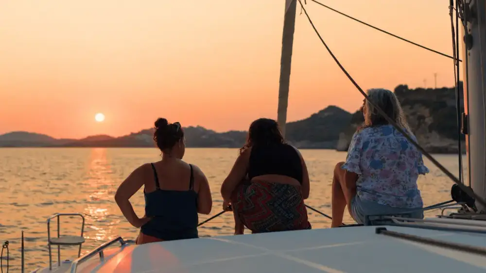 Three guests sitting together on the bow of a sailing catamaran, watching the sun set over the Mediterranean coastline as the sky glows orange during their yacht holiday.