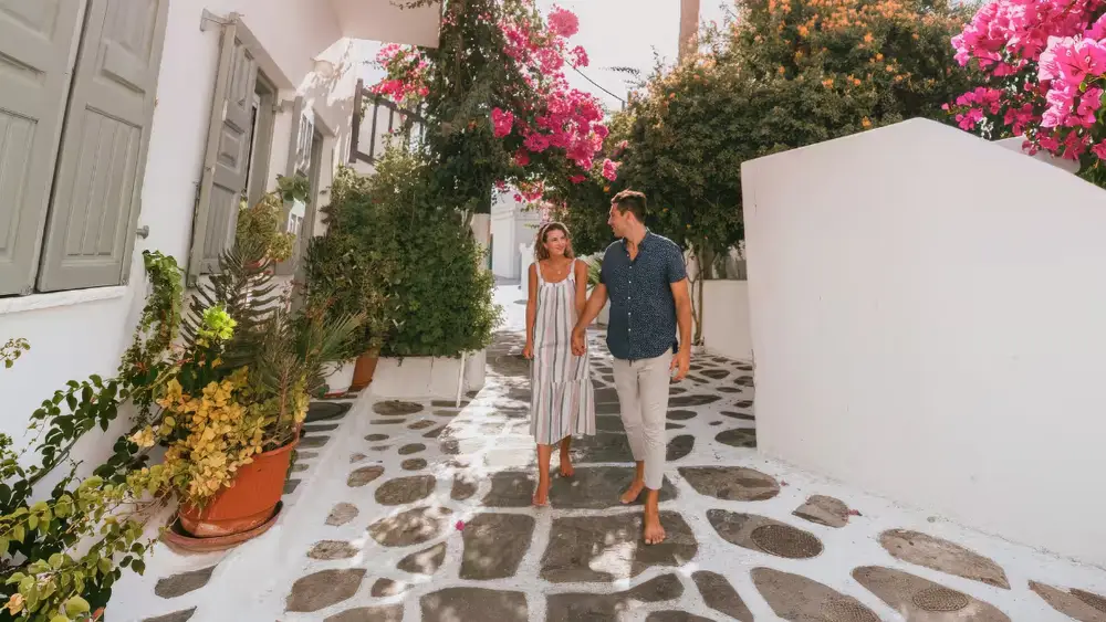 Couple walking through a whitewashed Cycladic village lined with colourful bougainvillea during a luxury yacht holiday through Paros and the Greek islands.