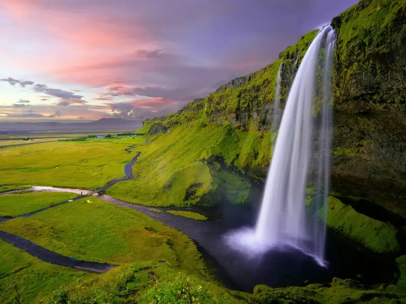 Seljalandsfoss waterfall on Iceland’s South Coast drops in a white curtain over green cliffs and open countryside under a colourful sunset sky. A classic Iceland nature scene that suits an article on the safest places to visit in Europe, reflecting Iceland’s long-standing reputation for peace, stability, and easy, low-stress travel for visitors exploring the country’s famous waterfalls and scenic routes.