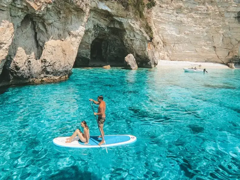 Guests paddleboarding over turquoise water near coastal caves on a Yacht Getaways sailing holiday, reflecting B Corp Certified responsible tourism in the Mediterranean.